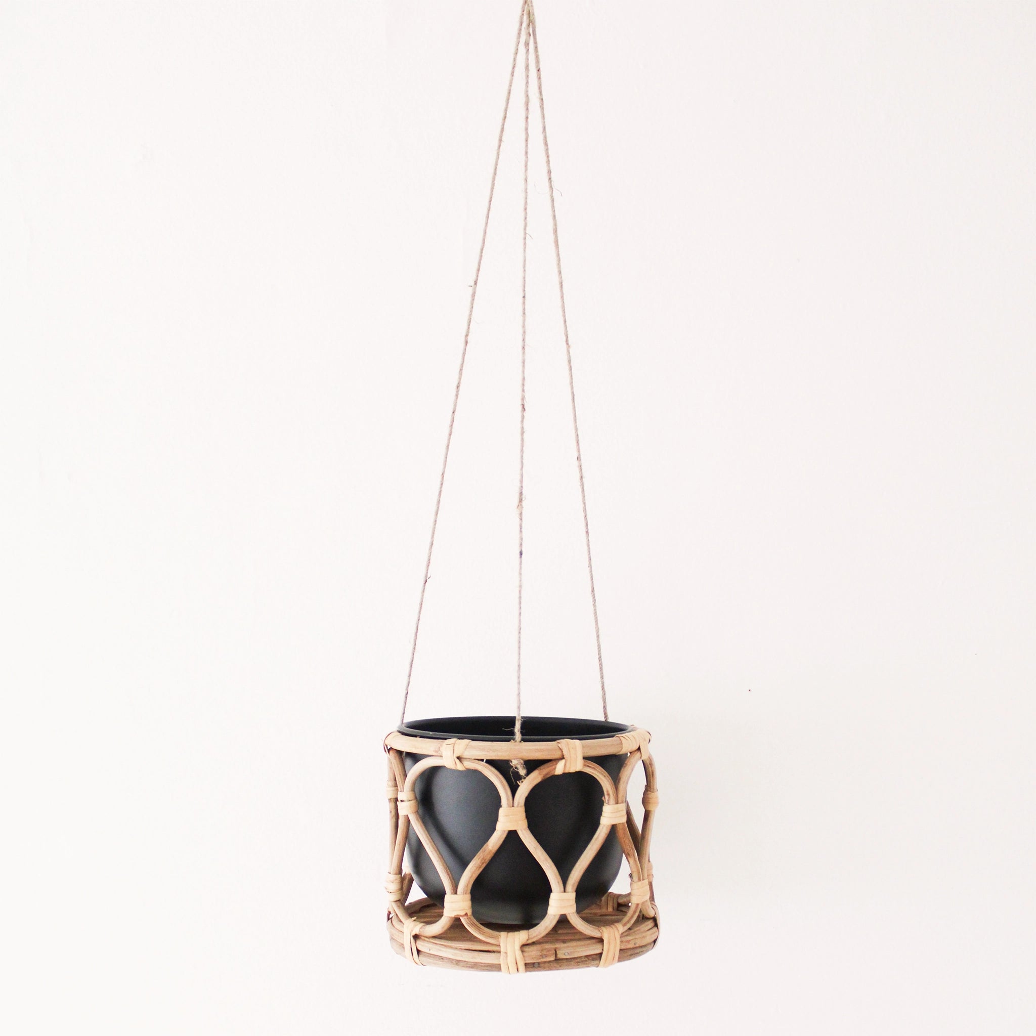 A hanging rattan planter with a black insert pot hanging in front of a white background. The rattan is bent into a rounded lattice pattern. The planter has a green trailing Pothos plant hanging inside of it.