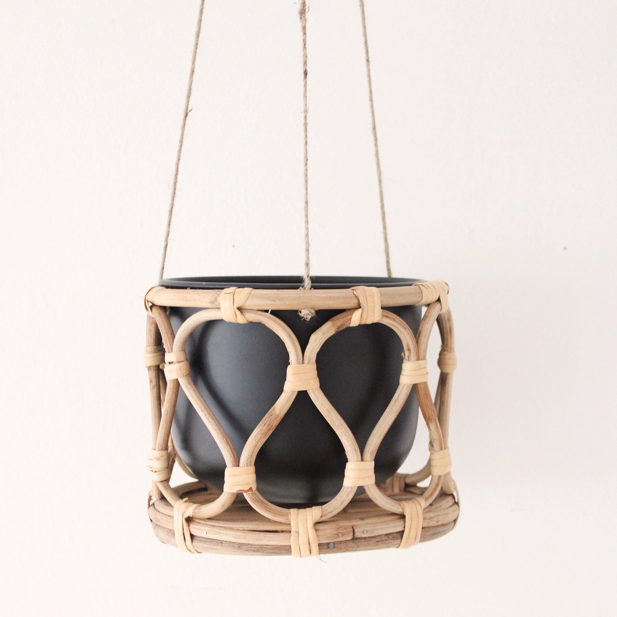 A hanging rattan planter with a black insert pot hanging in front of a white background. The rattan is bent into a rounded lattice pattern. Planter is hanging from 3 twine ropes that run off the edge of the photo.