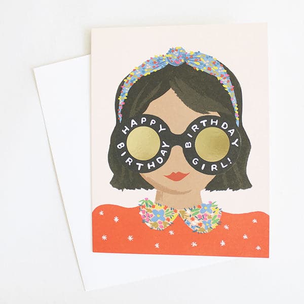 Photo of a greeting card on a white background. Card features the head and shoulders of a girl with short brown hair and tan skin with red lips on a pink background. She is wearing a blue floral headband and red shirt with floral collar. She has round black glasses that say &quot;happy birthday, birthday girl!&quot; on the frames. Gold foil fills in the inside of the glasses. Comes with a white envelope.
