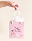 A cool toned pink paper gift bag with a graphic of a midcentury home in front of palm trees along with text that reads, "Warm Holiday Wishes" on the large size and "Merry Christmas" on the smaller one.