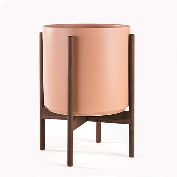 This 5 gallon, cylinder pot is a peachy terracotta color and sits within four spokes of a walnut wood plant stand, standing about 7 inches from the ground.