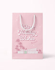 A cool toned pink paper gift bag with a graphic of a midcentury home in front of palm trees along with text that reads, "Warm Holiday Wishes" on the large size and "Merry Christmas" on the smaller one.