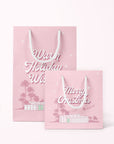 A cool toned pink paper gift bag with a graphic of a midcentury home in front of palm trees along with text that reads, "Warm Holiday Wishes" on the large size and "Merry Christmas" on the smaller one.
