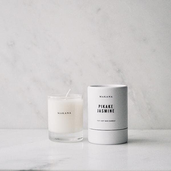 In front of a white and gray marbled wall is a round glass jar. Inside the jar is a white candle with a white wick in the center. On the front is black text that reads ‘Makana.’ To the right is a white round package. There is black text on the front that reads ‘Makana Pikake Jasmine.’ 
