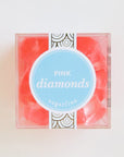 In front of a white background is a clear cube container. The container is filled with light pink diamond shaped gummies. In the middle is a bright blue round sticker with a white border. In the middle is white text that reads ‘pink diamonds.’
