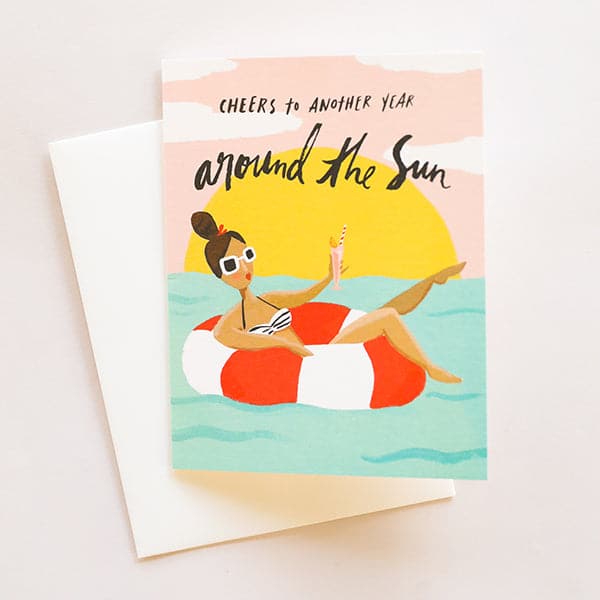 On top of a white envelope is a card. The card is a drawing of a blue ocean with a pink sky and yellow setting sun. In the pink sky is black text that reads ‘cheers to another year around the sun.’ In the water is a red and white striped inner tube with a girl laying inside. She is wearing white sunglasses and holding a pink drink. 