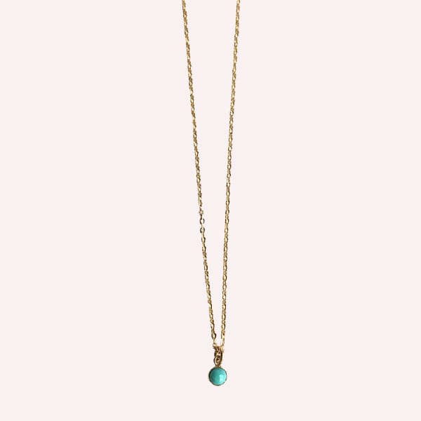 Hanging in front of a white background is a thin gold chain necklace. In the middle there is a small, round turquoise stone.