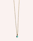 Hanging in front of a white background is a thin gold chain necklace. In the middle there is a small, round turquoise stone.