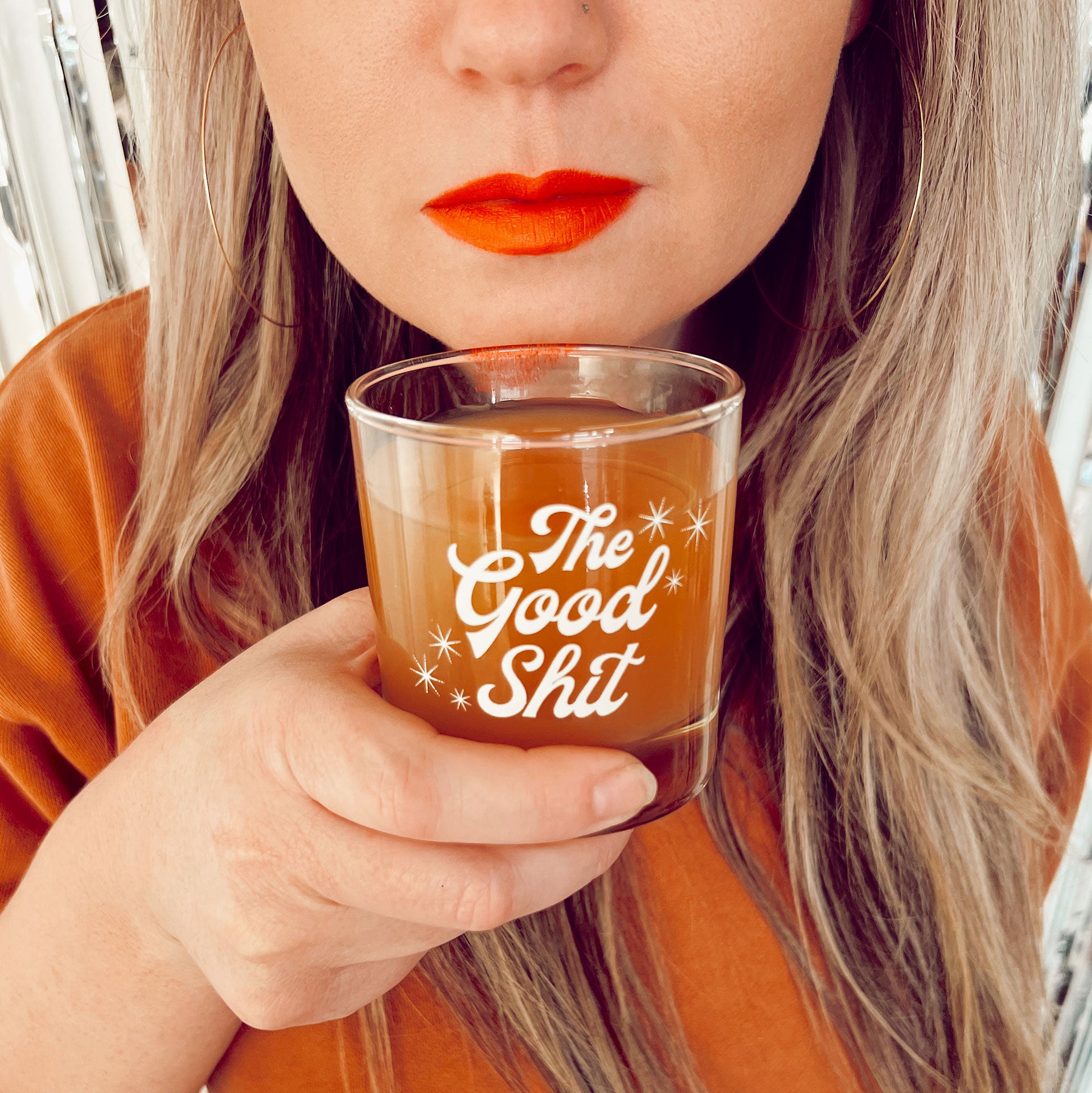 A photograph of a short glass tumbler with a thick bottom and &quot;The Good Shit&quot; printed across the center in white groovy cursive text.