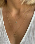 This is a picture of a woman’s neck and chest. She has short blonde hair and is wearing a white v-neck blouse. Around her neck is a thin gold chain necklace with a small turquoise bead in the middle. She is wearing a second thin gold chain necklace that falls below her collar bone. There is an oval shaped, gold pendent in the middle.