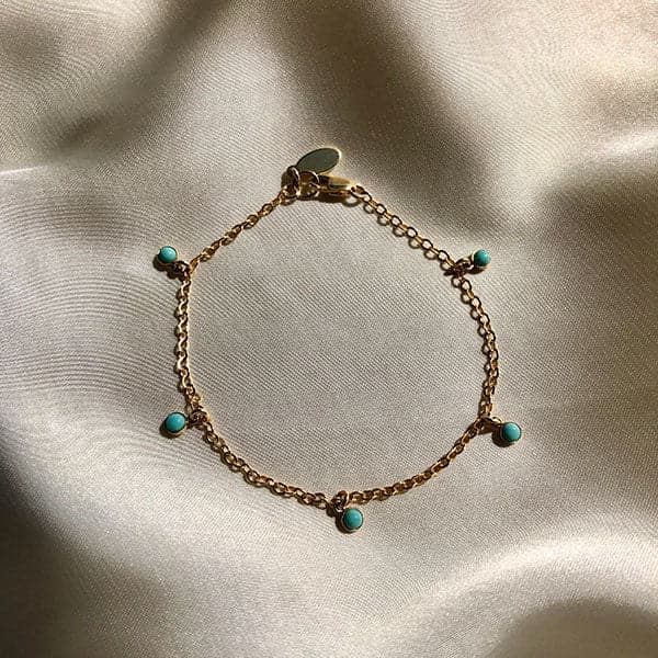 On top of a gray silky fabric is a thin gold chain bracelet. The bracelet has five circle turquoise stone charms.