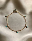 On top of a gray silky fabric is a thin gold chain bracelet. The bracelet has five circle turquoise stone charms.