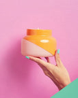 A hand is holding a squatty, round glass jar in front of a bright pink wall. The bottom corner of the background is a turquoise color. The model’s fingernails are painted the same turquoise color. The jar is split diagonally with the top half being a bright orange color and the bottom half being a soft pink color. The top of the jar is a lot narrower than the rest of the jar. There is a small flame inside the candle.