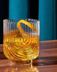 A clear glass tumbler with a gold base and an etched upside down arched design photographed here with a brown liquid and an orange peel garnish.