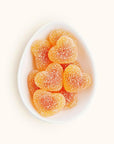 In front of a white background is a white egg shaped dish. The dish is filled with peach colored heart shaped gummies coated in white sugar.