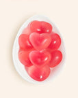 In front of a peachy background is the birds eye view of a white egg shaped dish. The dish is filled with bright pink heart shaped gummies.