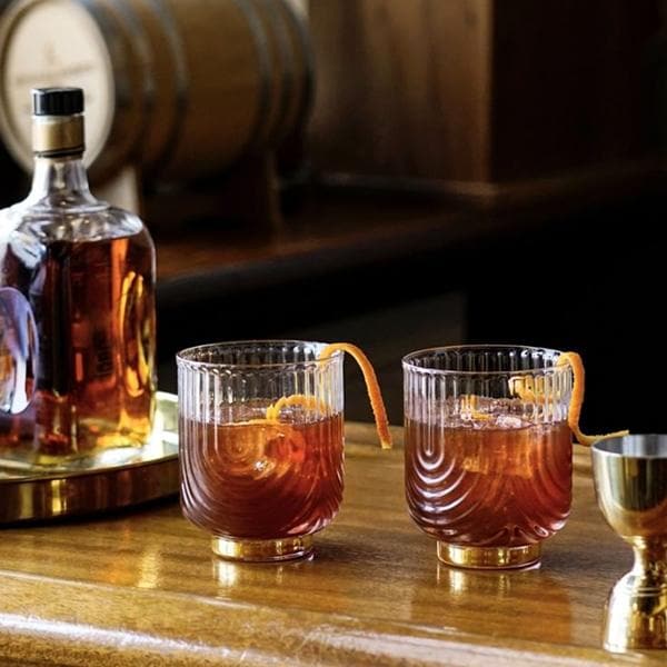 A clear glass tumbler with a gold base and an etched upside down arched design photographed here with a brown liquid and an orange peel garnish.