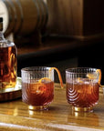 A clear glass tumbler with a gold base and an etched upside down arched design photographed here with a brown liquid and an orange peel garnish.
