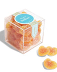 In front of a white background is a clear plastic cube container. The container is filled with peach colored heart shaped gummies coated in white sugar. On the top of the container is a bright blue circle sticker with white text that reads ‘peach Bellini.’ There are three gummies sitting outside of the container.
