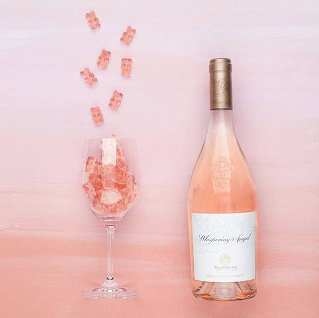 In front of a pink background is a glass bottle of light pink rosé. It has a gold top. To the left is a clear wine glass. The glass is filled with light pink gummy bears. 