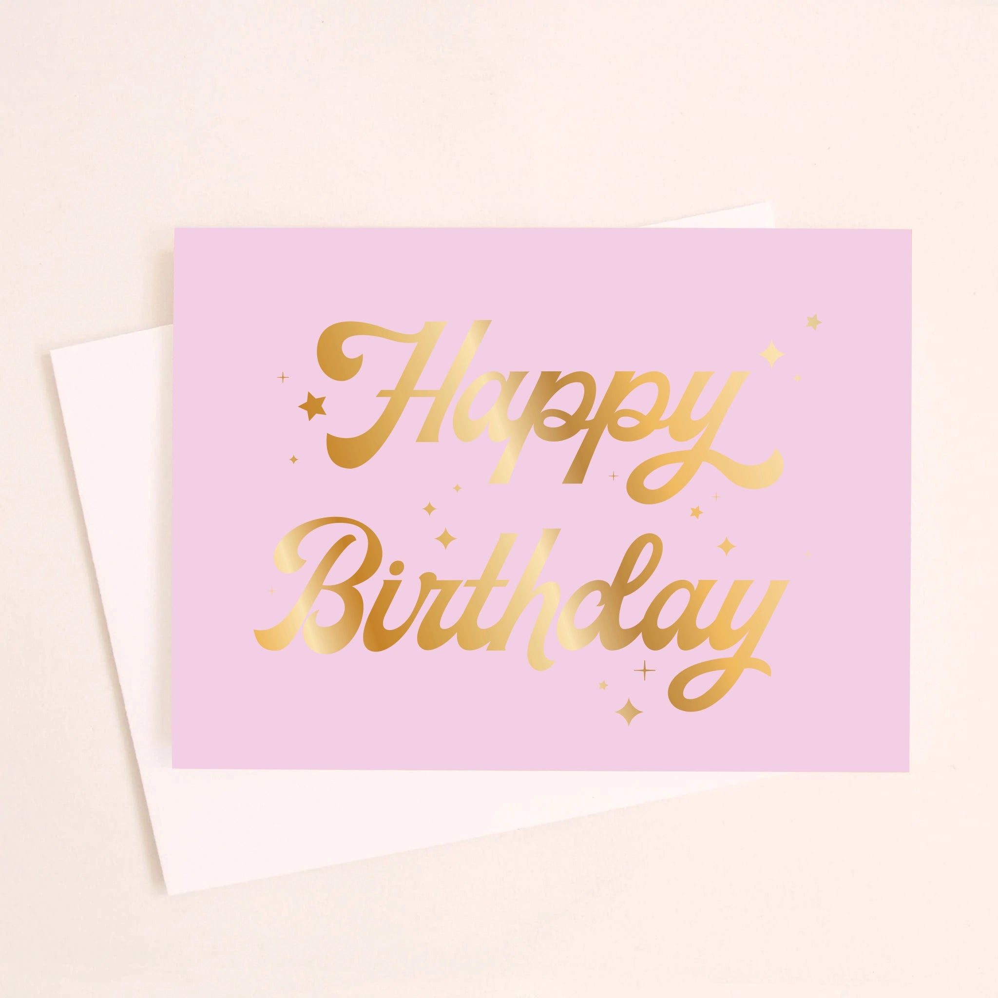 On a cream background is a lavender card with gold foiled cursive text that reads, &quot;Happy Birthday&quot; along with a white envelope.