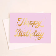 On a cream background is a lavender card with gold foiled cursive text that reads, "Happy Birthday" along with a white envelope.