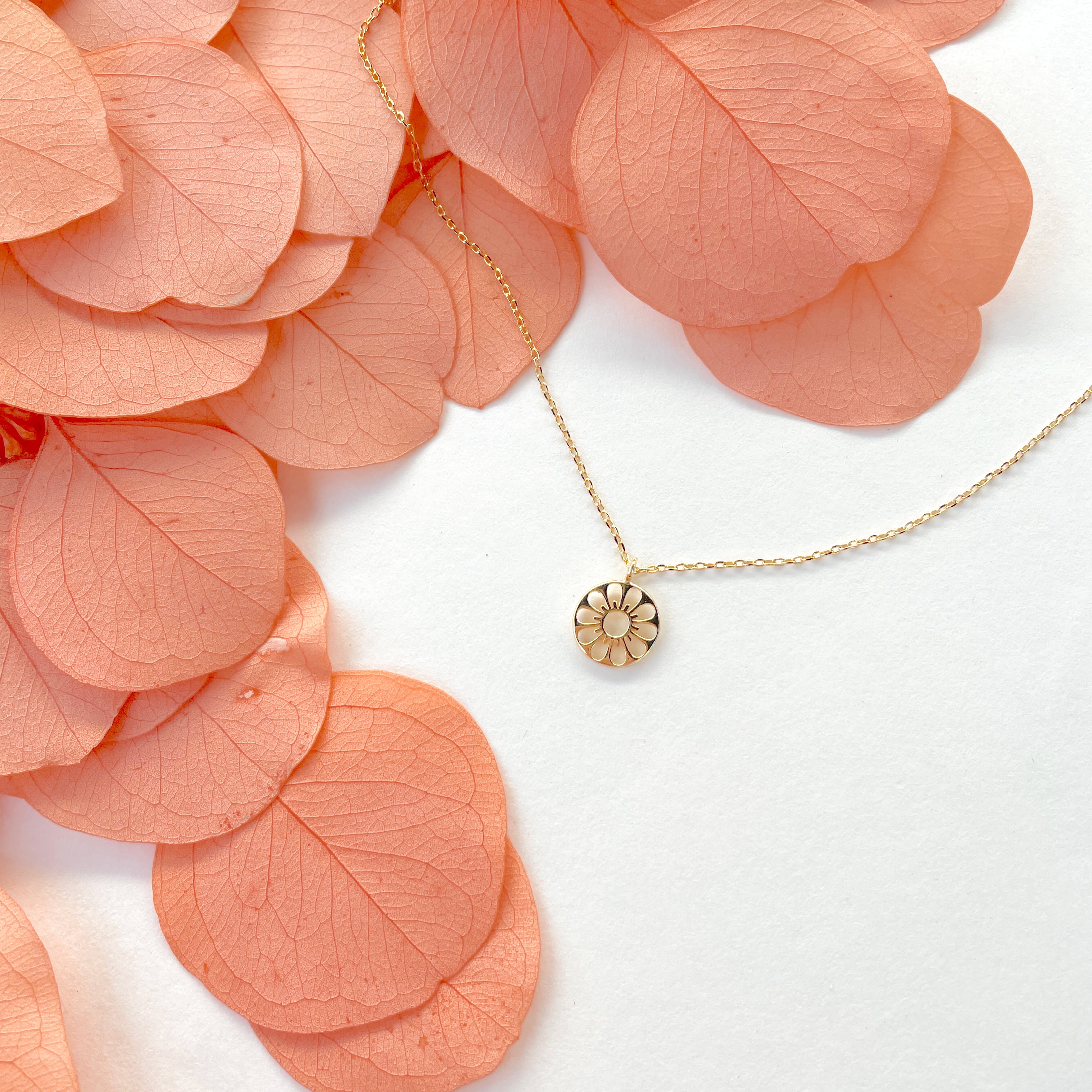 A  dainty gold chain necklace with a gold circle pendant in the center with a cut out of a daisy flower, photographed next to pink dried florals.
