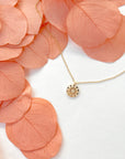 A dainty gold chain necklace with a gold circle pendant in the center with a cut out of a daisy flower, photographed next to pink dried florals.