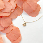 A dainty gold chain necklace with a gold circle pendant in the center with a cut out of a daisy flower, photographed next to pink dried florals.
