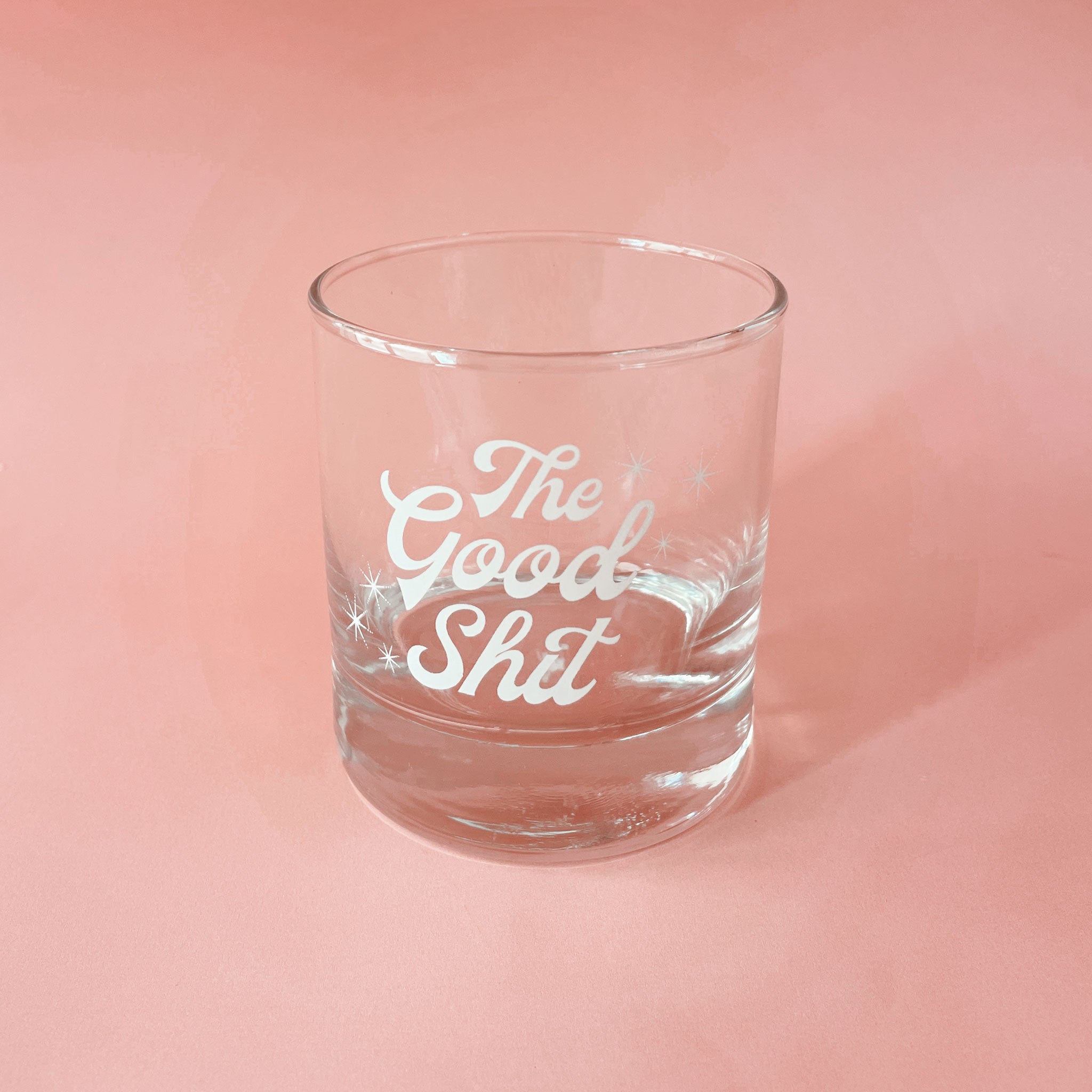 Against a peachy pink background is a photograph of a short glass tumbler with a thick bottom and &quot;The Good Shit&quot; printed across the center in white groovy cursive text.
