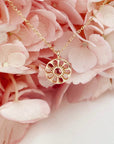 A dainty gold chain necklace with a gold circle pendant in the center with a cut out of a daisy flower, photographed in front of pink hydrangeas.