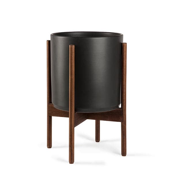 This cylinder pot is a solid black and sits within four spokes of a dark wood plant stand, standing about 7 inches from the ground.