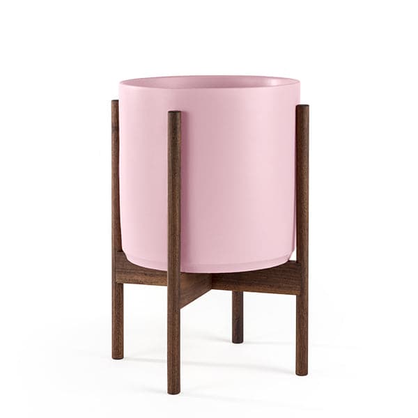 This cylinder pot is a baby pink and sits within four spokes of a walnut wood plant stand, standing about 7 inches from the ground.