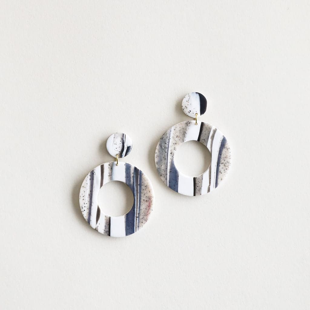The Mollye earrings that feature a duel circle design. The top circle is smaller and solid while the lower circle is larger and open in the center. This earring features a straight post backing and comes in a variety of colors. This color way is sandstone and white.