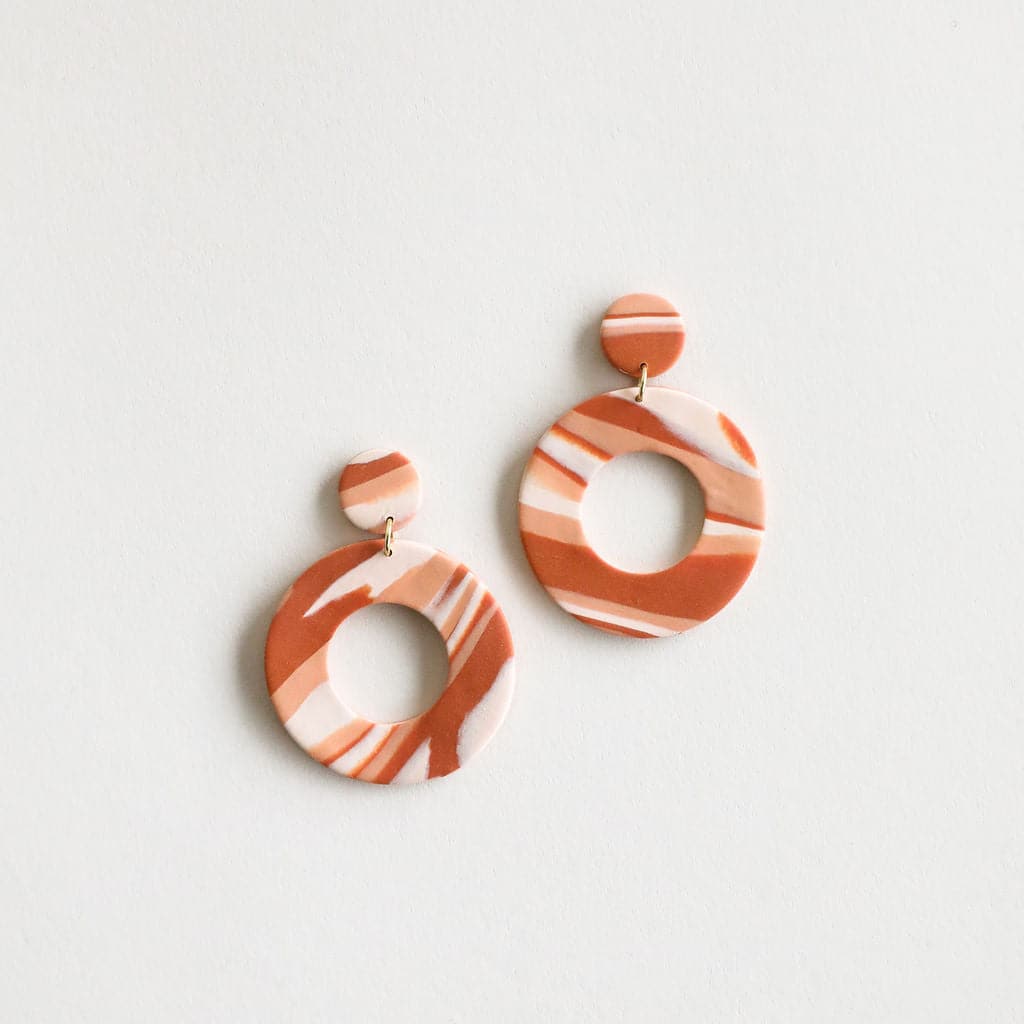 The Mollye earrings that feature a duel circle design. The top circle is smaller and solid while the lower circle is larger and open in the center. This earring features a straight post backing and comes in a variety of colors. This color way features rust, salmon and cream tones. 