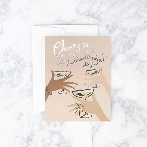 Neutral toned card reading &#39;Cheers to the bride to be&#39; in silver foil. Below three hands cheer glasses of champagne. 