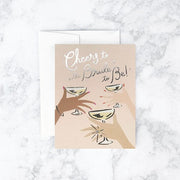 Neutral toned card reading 'Cheers to the bride to be' in silver foil. Below three hands cheer glasses of champagne. 