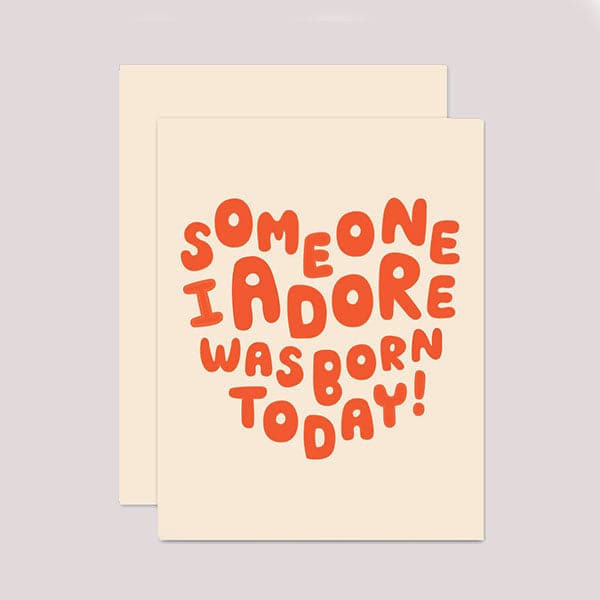 An ivory card with matching envelope. On the front of the card is red bubble text in the shape of a heart saying "Someone I Adore Was Born Today."