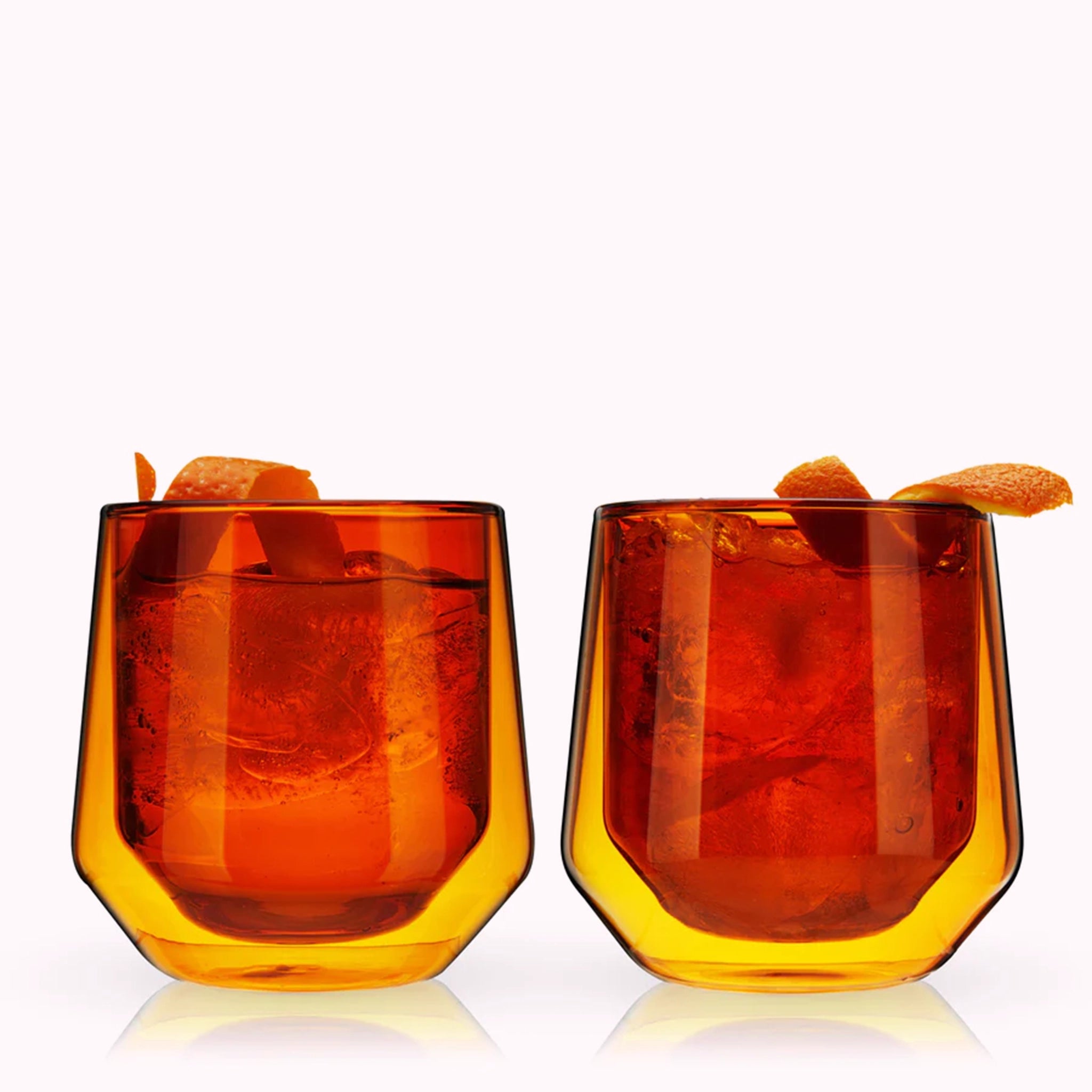 A set of two double walled glass tumblers in a vibrant amber color photographed here in front of a white background with orange peel garnishes.