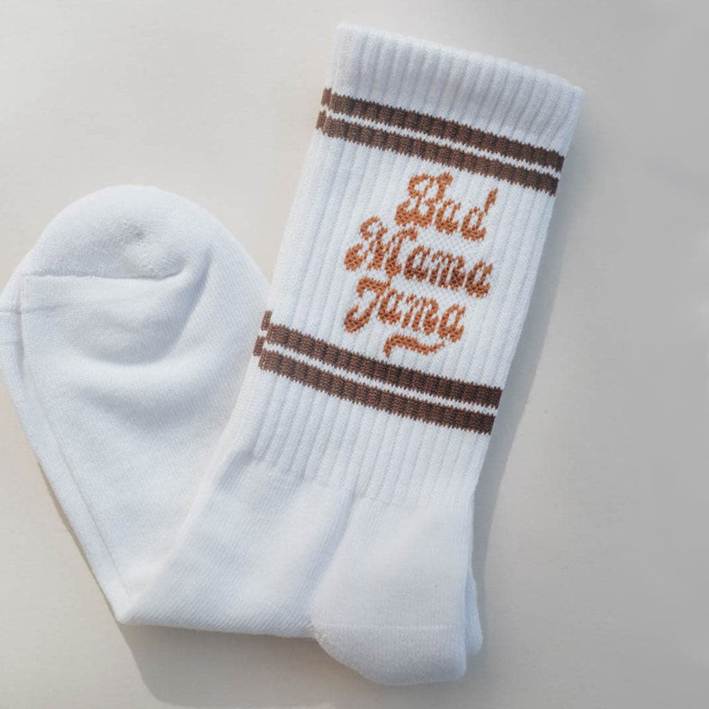 Pair of ribbed white tube socks that reads &#39;Bad Mama Mama&#39; in burnt orange lettering. Two brown stripes fall above and below the text giving them a retro look. 