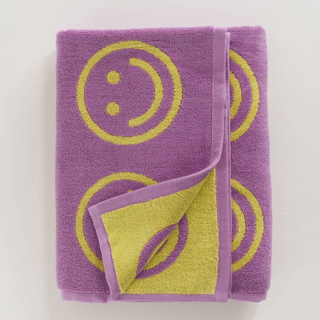 Light purple cotton towel covered in neon yellow classic smiley faces. The corner of the towel is pealed forward revealing an inverted version of the pattern on the backside, yellow cloth with purple smiley faces. 
