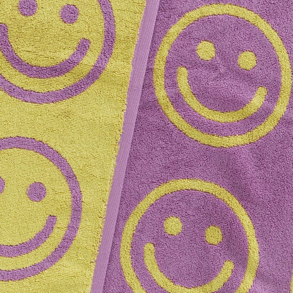 Both sides of the towel positioned side by side one another. To the left is the yellow towel with purple smiley faces and to the right is the purple towel with yellow smiley faces. 