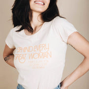 Woman modeling an ivory white t-shirt with peach retro lettering, "Behind Every Great Woman, There's a Shit Ton of Other Dope Ass Women."