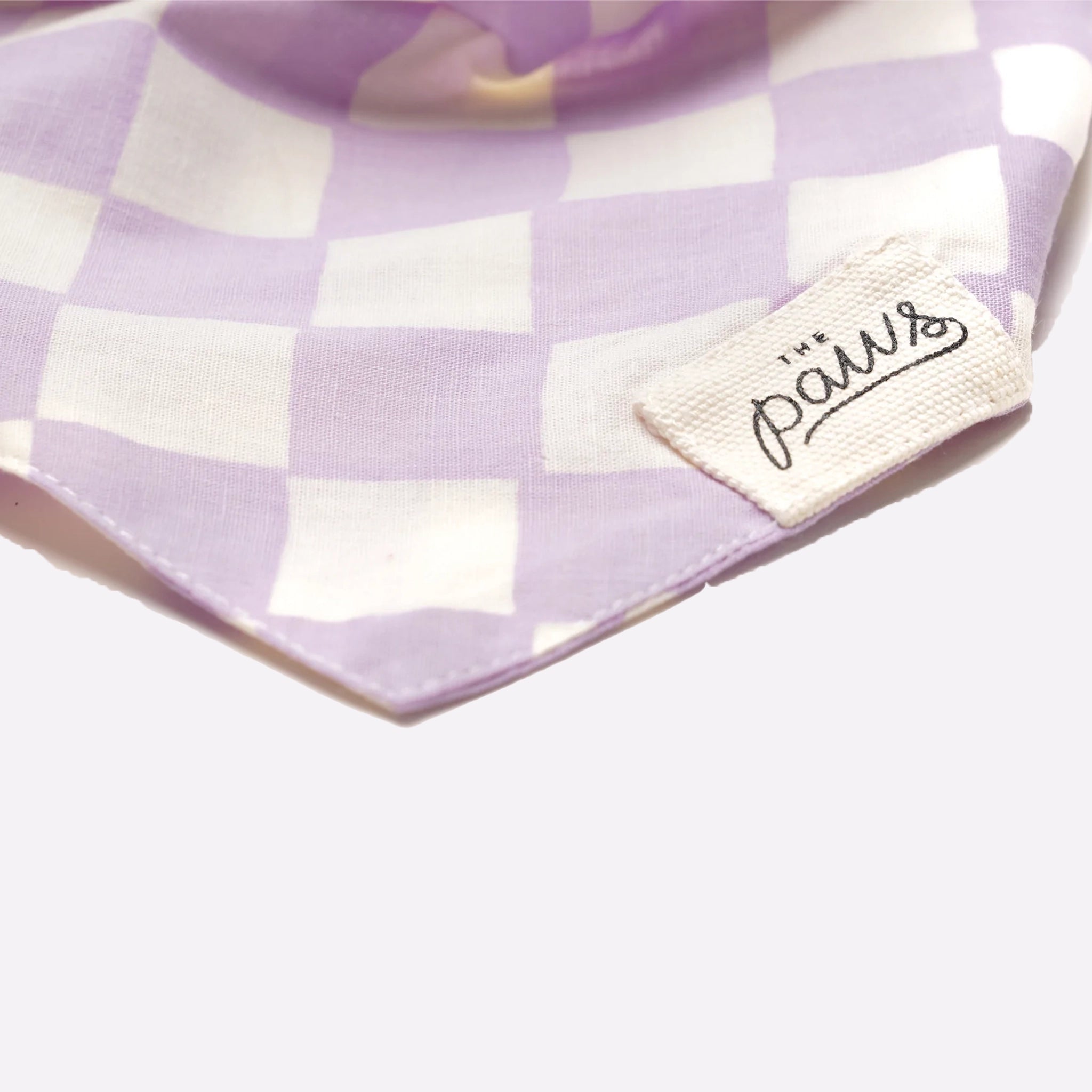 A lavender and white checkered pet bandana with a tie back and a small label on the front that reads the brand's name, "The Paws".