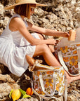 Photographed in front of a yellow background is a mini cooler with a cream adjustable strap and 70's style yellow and orange floral print photographed with a model at the beach.