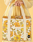 Photographed in front of a yellow background is a picnic bag with cream straps and 70's style yellow and orange floral print.