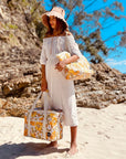 Photographed in front of a yellow background is a picnic bag with cream straps and 70's style yellow and orange floral print held by a model on a beach.