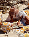 A 70's style yellow and orange floral picnic mat with cream fringe detailing around the edges photographed with coolers and beach bags in the same print with a model laying on it reading a book.