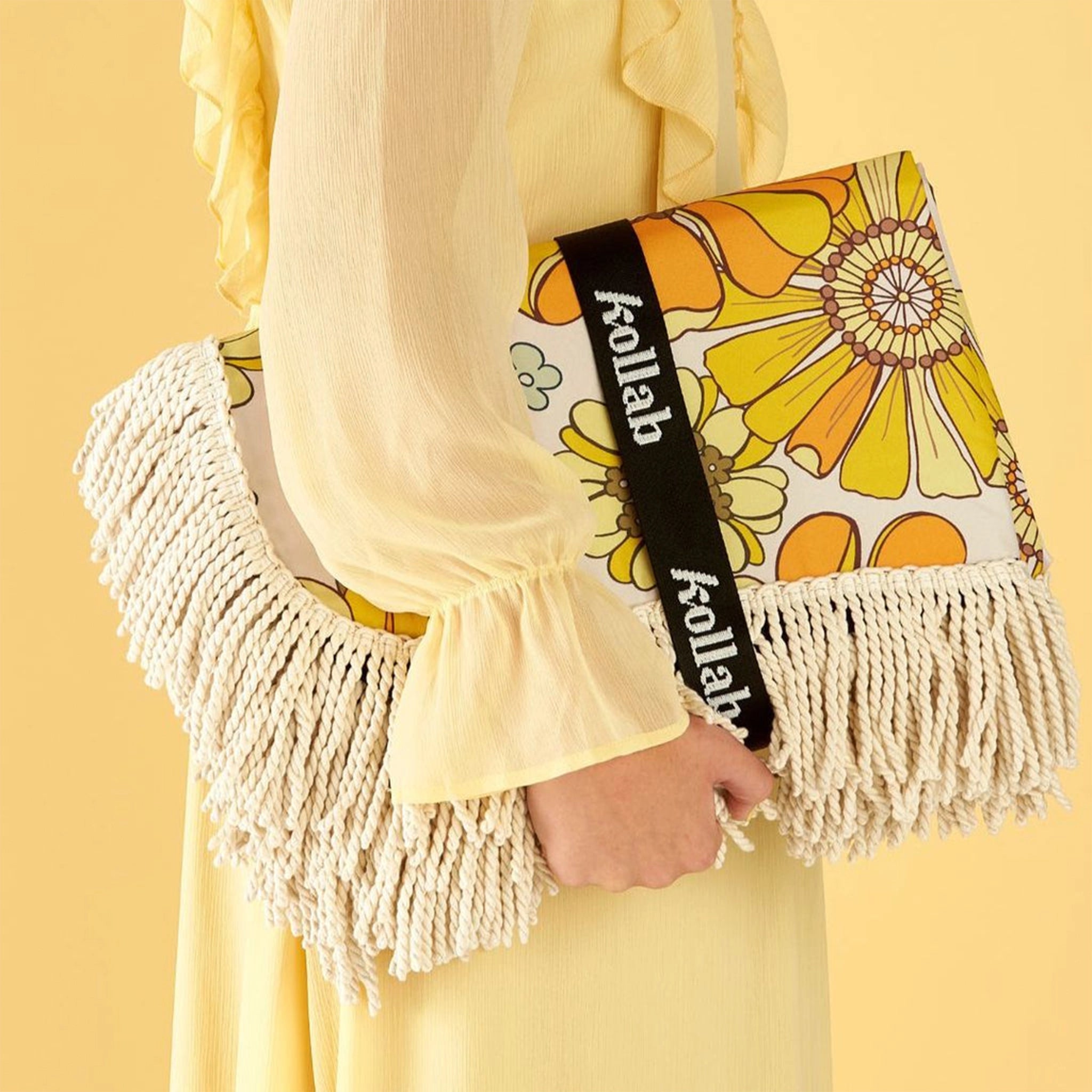 A 70&#39;s style yellow and orange floral picnic mat with cream fringe detailing around the edges photographed with a model holding it in front of a light yellow background. 