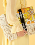 A 70's style yellow and orange floral picnic mat with cream fringe detailing around the edges photographed with a model holding it in front of a light yellow background.